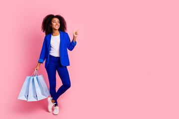 Full length photo of cheerful woman wear blue jacket hold shopping bags look directing empty space isolated on pink color background