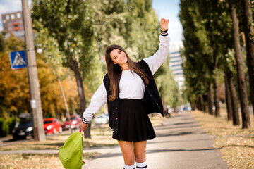 Photo of optimistic girlish student with straight hairstyle dressed stylish jacket hold bag raising arm up in sunny weather outdoors