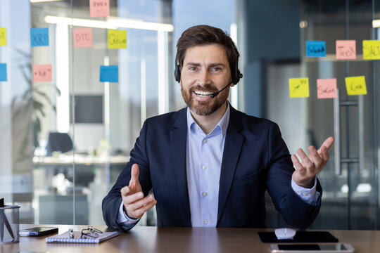 Cheerful businessman wearing headset and talking during a video conference in a modern office. The background features colorful sticky notes on a glass wall. Concept of communication