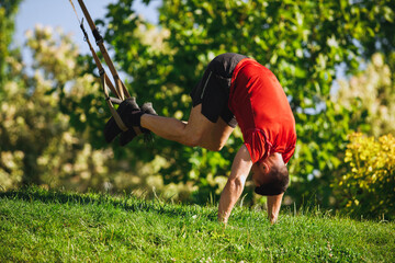 Fit athlete, muscular man in sportswear training outdoors in park, performing an intense TRX pike workout on a grassy ground. Concept of sport, body and health care, active lifestyle