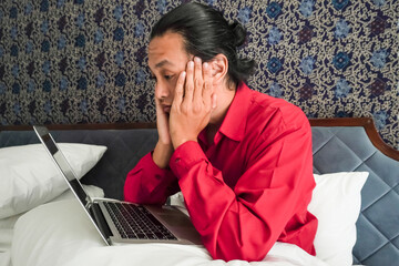 Bored long-haired young asian man in red long-sleeve shirt is not interested while working remotely...