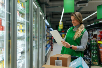 Beautiful woman working in a supermarket. Female assistant using a digital tablet for taking inventory of products in display rack in a grocery store.