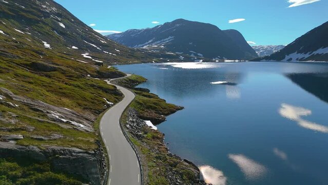 A mountain road winds by a tranquil lake with snowy peaks offering a breathtaking view of the serene landscape. Langvatnet, Geiranger, Norway