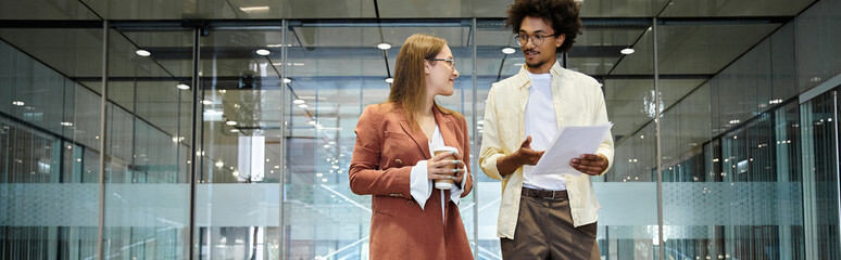 Two colleagues chat in a modern office lobby, one holding a coffee cup and the other reviewing a...