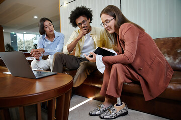Three colleagues, including a woman with a prosthetic leg, discuss a project on a tablet while sitting on a couch in a modern office lobby.