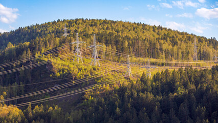 Power lines pylons with electric wires in mountainous area covered with coniferous forest. High voltage power transmission towers