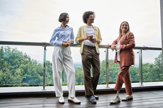 Three colleagues, diverse in race and ability, enjoy a casual conversation on an office rooftop terrace.