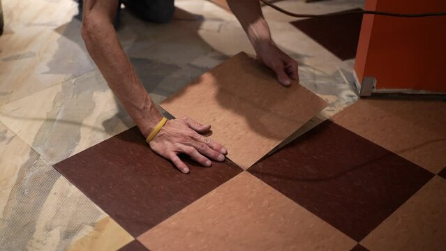 With glue on the floor, two hands hold a VCT tile and carefully move it into place, pressing it down into the glue.