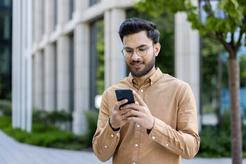 Young businessman using smartphone outside office building, focused on device. Casual attire,...