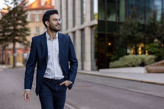 Confident smiling businessman wearing blazer and glasses walking in urban city street. Professional young man enjoying his day. Concept of success, confidence, business attire. - Powered by Adobe