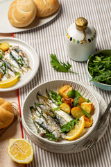Table with Mediterranean style lunch, sardine and baked baby potato salad with arugula. Oven baked italian fish sardoncini or sardines, with olive oil, lemon and parsley. Vertical image.