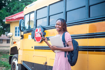 youthful pretty African adult female student using mobile technology while holding laptop and standing close to a school bus