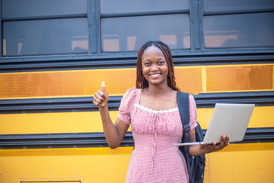 youthful pretty African adult female student using mobile technology while holding laptop and standing close to a school bus