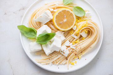 Plate of linguine pasta with feta cheese, lemon zest and basil, horizontal shot on a beige marble surface, middle closeup