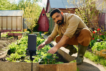Bearded mature farmer looking at online audience on smartphone screen while bending over flowerbed...