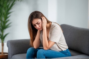 Fototapeta premium A woman sits on a couch at home suffering from a headache or migraine with copy space.