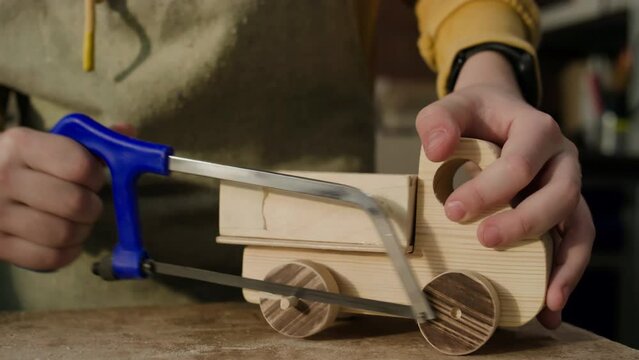 Close up view of hands of little boy using hacksaw while cutting wheels of wooden toy truck with help of woodworking teacher