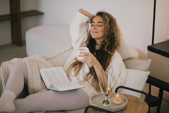 Young woman reading book and drinking tea, sitting on sofa in a cozy living room.