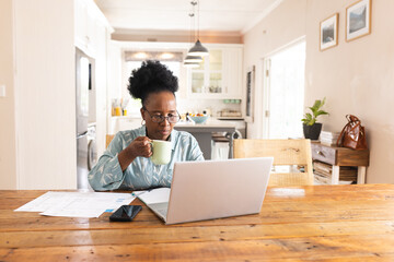 Busy senior african american woman with documents on table using laptop at home