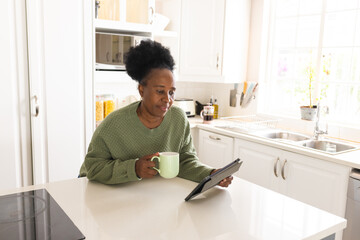 Happy senior african american woman drinking coffee and using tablet in kitchen at home