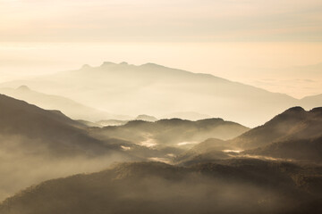 A hazy, golden sunrise illuminates a series of rolling mountain ranges, their peaks fading into a soft, misty background. 