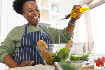 Happy senior african american woman making salad and pouring oil in kitchen at home