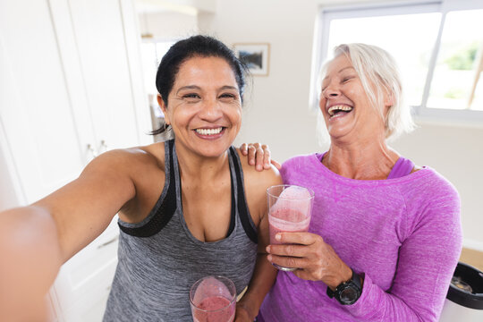Two happy diverse senior female friends drinking fruit smoothies taking selfie in kitchen