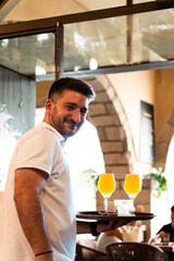 Waiter in white polo shirt with two beers on a tray held with one hand while smiling. Vertical