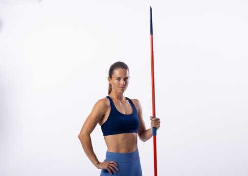 Confident young Caucasian female athlete with javelin poses on white, space for text