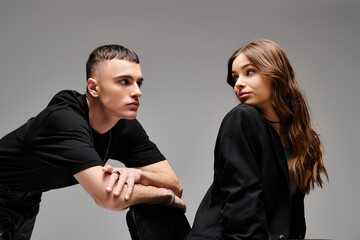 A young man and woman sit side by side, sharing a moment of closeness and connection in a studio with a grey background.