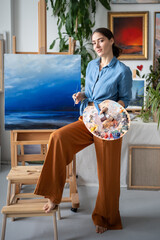 Contented creative woman artist holds colour palette, brush standing near wooden easel with...