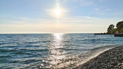 A wave rolls over the sea pebble beach. Sun glare on the water. There are trees growing on the...