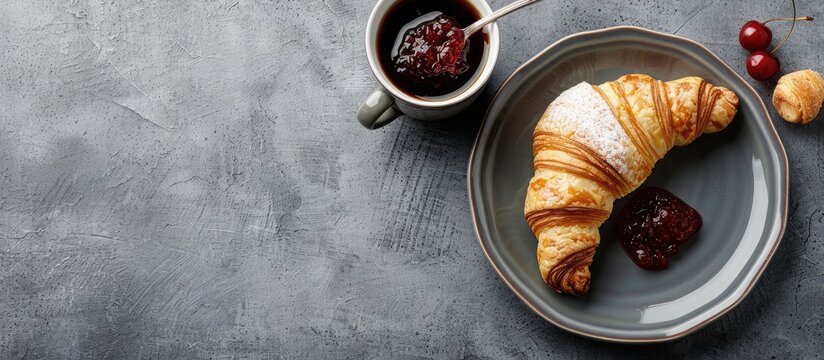 Delicious sweet breakfast with pastries: a fresh French croissant with cherry jam on a grey plate, a cup of coffee nearby, and a teaspoon spreading jam, all displayed on a gray background with copy
