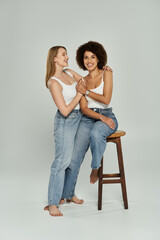 Two women in jeans and tank tops pose together in front of a grey background.