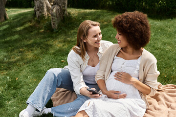 Fototapeta premium Two women, one pregnant, share a tender moment while sitting on a blanket in a grassy park.