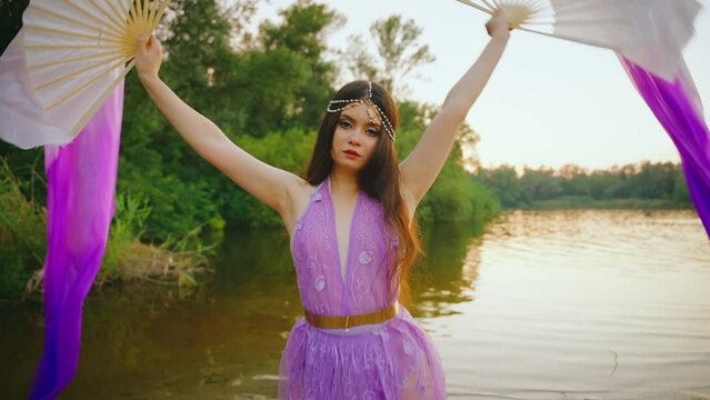 Fantasy fairy girl waving hands holding two dancing fans, long silk fabric flies in wind slow motion belly dance, purple dress. Woman goddess elf queen. river lake forest green trees, summer nature 4k