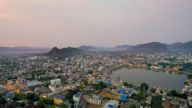 Aerial drone orbit shot over hindu holy city of pushkar with lake bhrama temple visible in city lights and aravalli hills covered in fog in distance showing cityscape in Rajasthan India