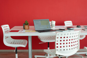 Modern office workspace with red wall in background, featuring laptop, coffee cup, and other office essentials on white table