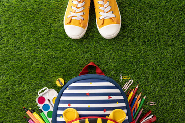 Top view of back to school supplies with backpack and yellow sneakers placed on green grass