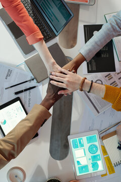 Close-up of team members stacking hands over desk in collaborative office meeting. Diverse hands symbolizing unity and teamwork with digital devices and documents in background
