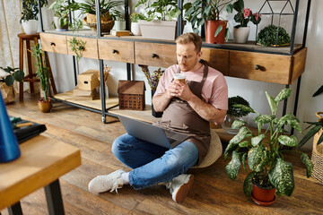 A man with a business shirt sits on floor, engrossed in laptop, surrounded by potted plants in a...
