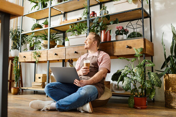 A stylish man sits on the floor, focused on his laptop at his small plant shop. He is busy managing...