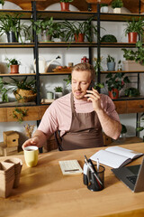 A man sits at a desk in a plant shop, talking on a cell phone, showcasing determination towards his small business success.