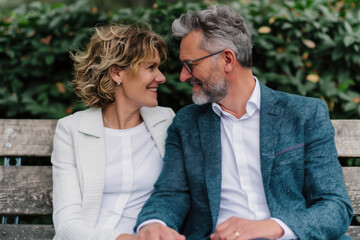 Obraz premium Happy elderly couple sitting on park bench, looking at each other lovingly, surrounded by lush greenery, enjoying peaceful moment together.