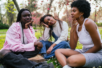 Fototapeta premium Three young African women sitting thoughtfully on the grass in a park, looking at the camera.
