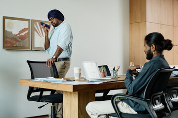 Skilled boss having conversation via smartphone while his young Indian subordinate sitting at table and waiting for instructions