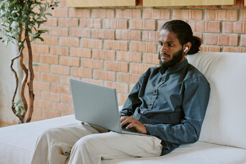 Young Indian male freelancer sitting on velvet sofa while surfing Internet and listening to music