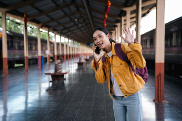 Energetic young woman at bustling train platform, chatting on phone and waving with enthusiasm, eager for a new journey
