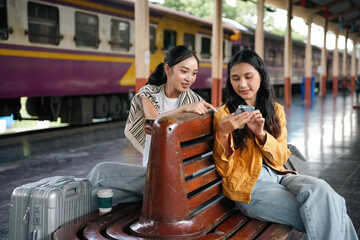 Two pals are seated on a train station bench, occupied with a smartphone while waiting for their train to arrive