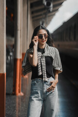 Young woman waits on a busy train platform, adjusting her sunglasses with confidence and style,...